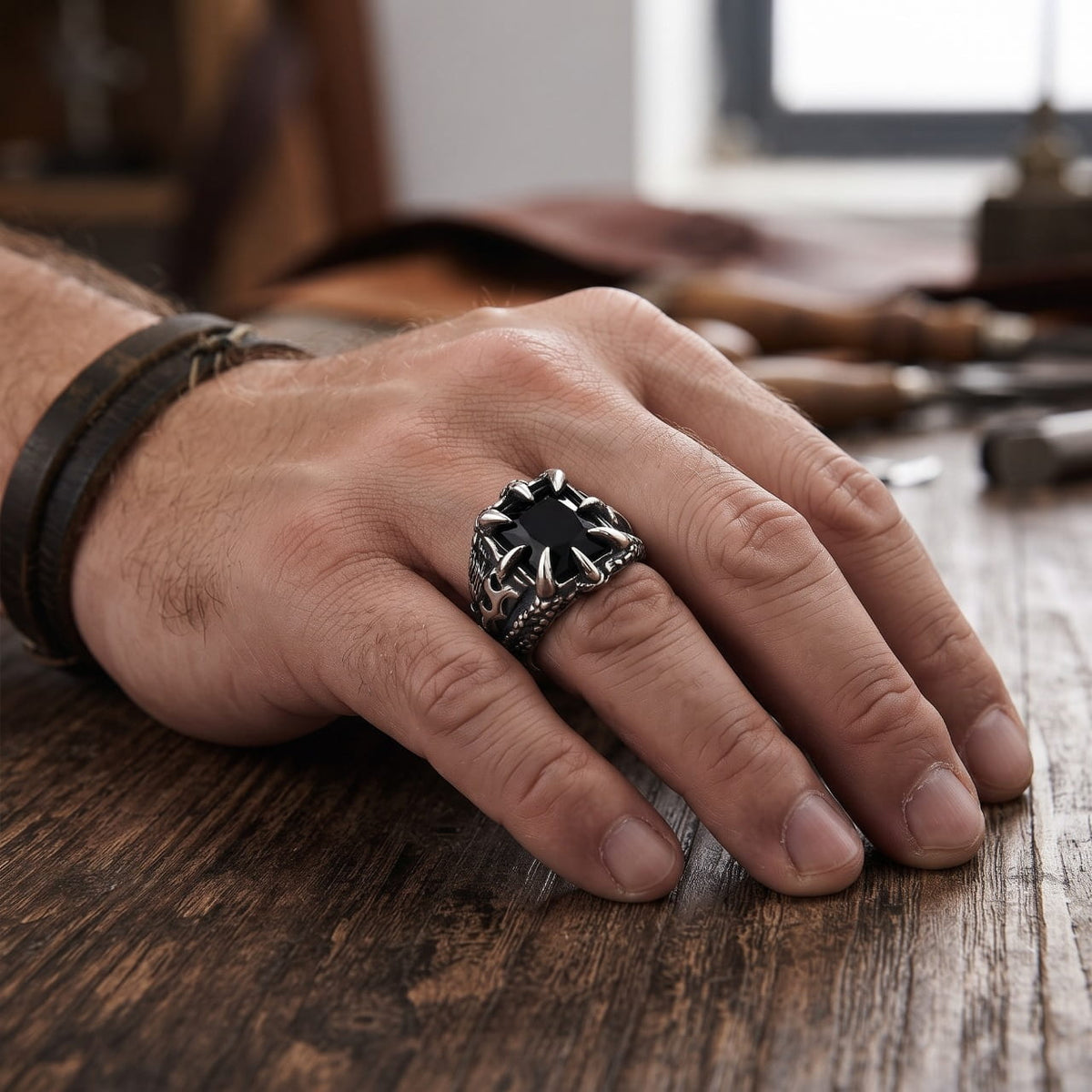 Bague en acier à griffes de dragon enserrant une pierre noire portée à l'annulaire d'une main d'homme, bracelet de cuir au poignet