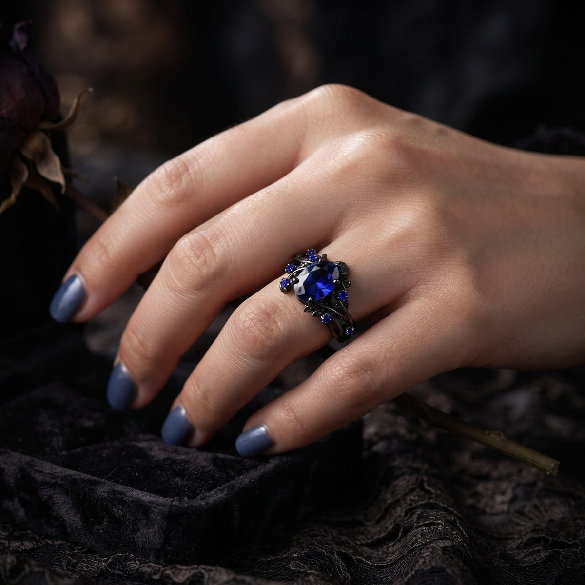 Bague en acier noir sertie d'un zircon bleu portée à l'annulaire d'une main féminine posée sur du velours sombre