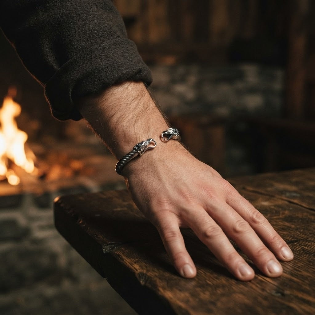 Bracelet viking jonc en acier torsadé à têtes de corbeau, porté au poignet d'un homme, main posée sur une table en chêne près du feu