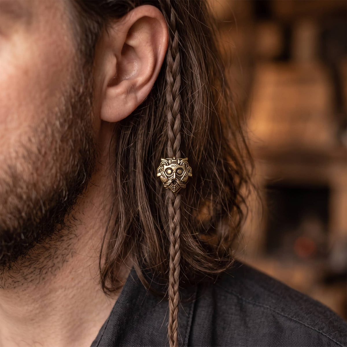 Perle viking en alliage bronze patiné, en forme de masque barbu aux yeux ronds et à la moustache tombante, portée sur une fine tresse de cheveux