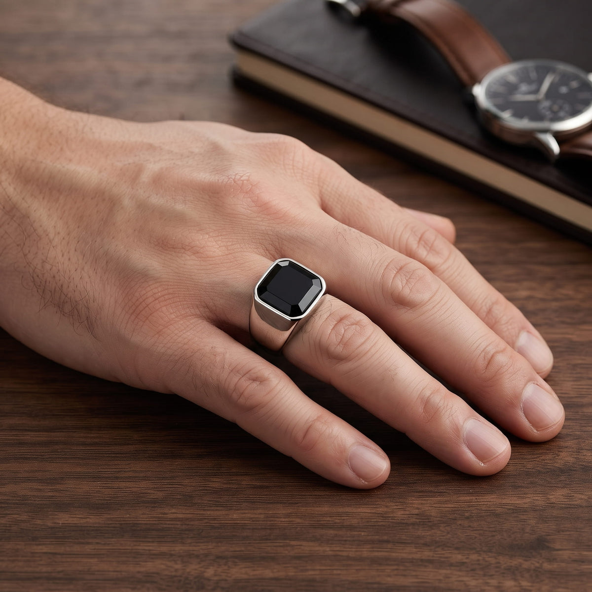 Bague en acier poli sertie d'une agate noire carrée portée à l'annulaire d'une main d'homme, montre en cuir sur le bureau
